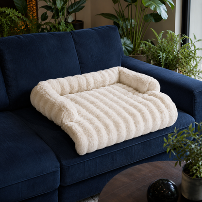 A plush cream-colored pet bed with a ribbed texture and raised corners, placed on a dark blue sofa in a living room setting with potted plants in the background.
