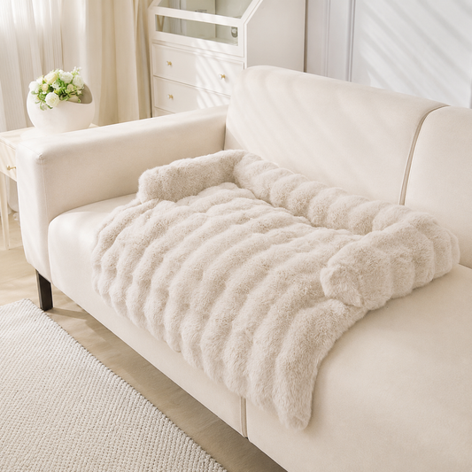 A cream-colored, plush faux fur pet bed with a raised, ribbed backrest, styled on a matching cream sofa in a bright, modern living room.